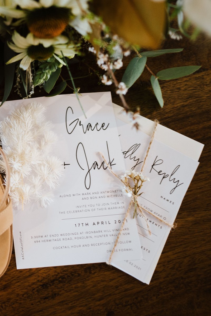 Linear wedding invitation and RSVP card with handwritten style names, tied with twine and dried flowers near bridal sandals